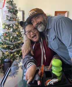 Christmas tree in the background. Mikelle in her wheelchair is being hugged by her brother who has a beard and ballcap on.