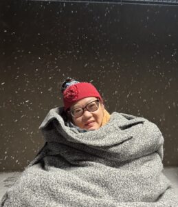 Mikelle is in her wheelchair bundled up with a gray blanket at night with the snow falling around her.