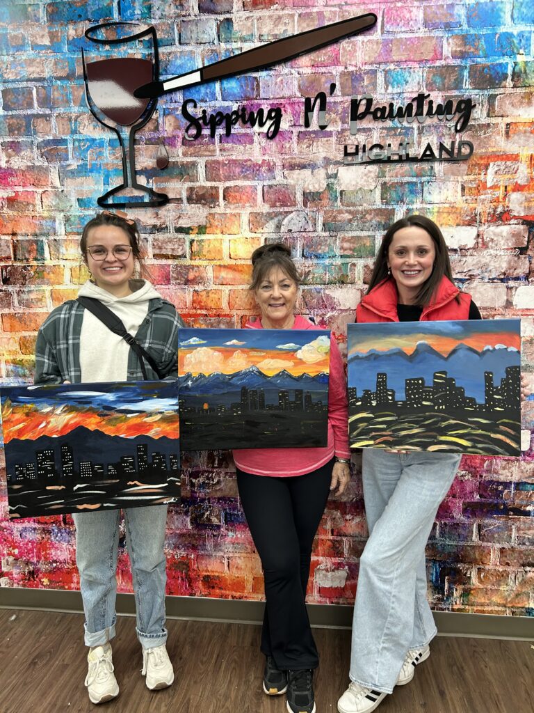 Three women including Maryann, Katherine and Liz holding up their paintings of the Colorado mountains and the skyline of Denver at sunset.
